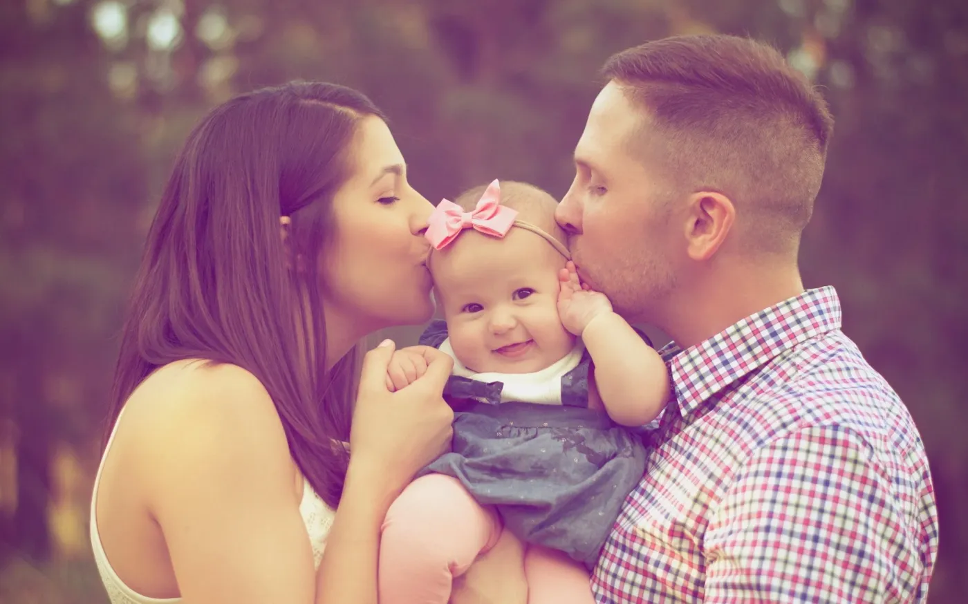 A man and woman kiss a baby while standing together in a sunny field, surrounded by green grass and wildflowers.