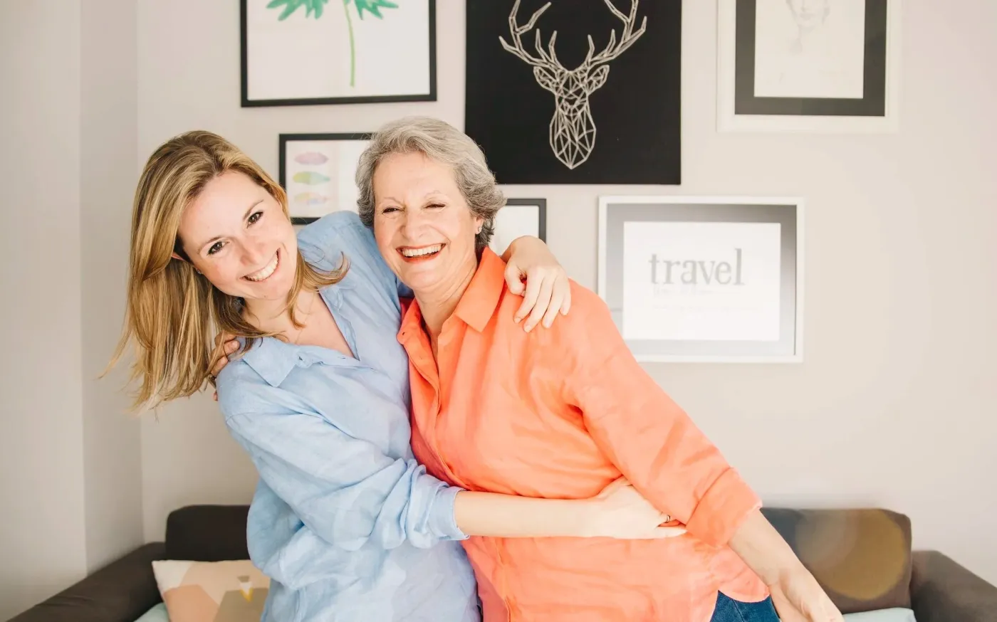 Two women embrace warmly in front of a wall adorned with various pictures.