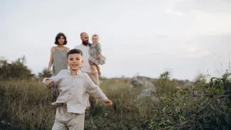 A family joyfully running together through a lush green field under a clear blue sky.