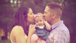 A man and woman kiss a baby while standing together in a sunny field, surrounded by green grass and wildflowers.
