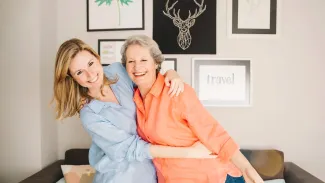 Two women embrace warmly in front of a wall adorned with various pictures.